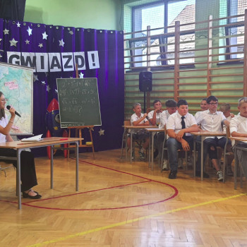 Uczniowie podczas przedstawienia, dziewczyna siedzi przy stoliku po prawej stoi tablica