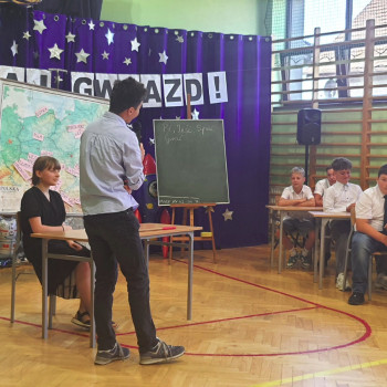 Uczniowie podczas przedstawienia, dziewczyna siedzi przy biurku, chłopak stoi przy tablicy