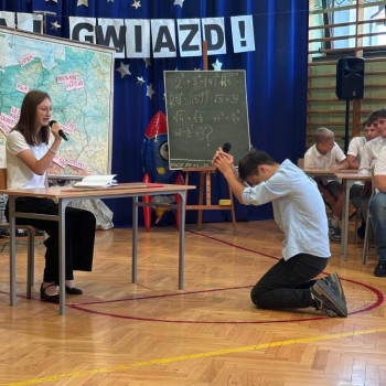 Uczniowie podczas przedstawienia, uczeń klęczy przed biurkiem nauczycielki